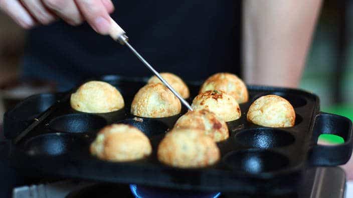 A view of a person checking if the octopus balls are done in a Takoyaki pan with the help of a stick