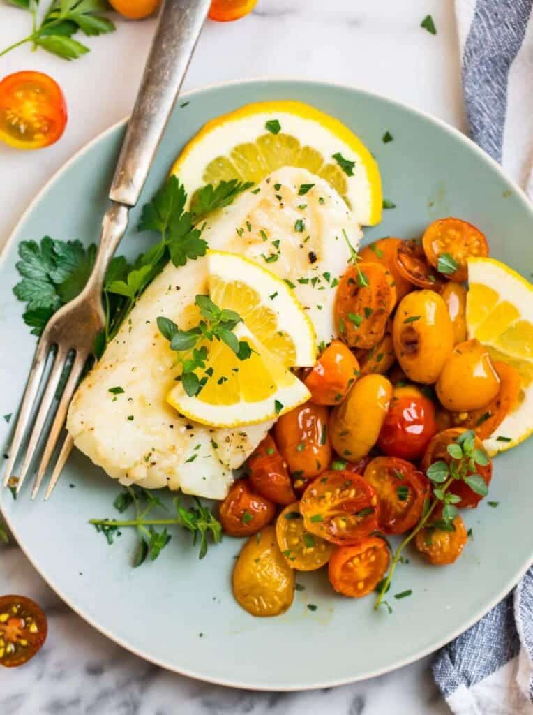 cod fish served in a plate with a fork cilantro fried tomatoes and lemon wedges