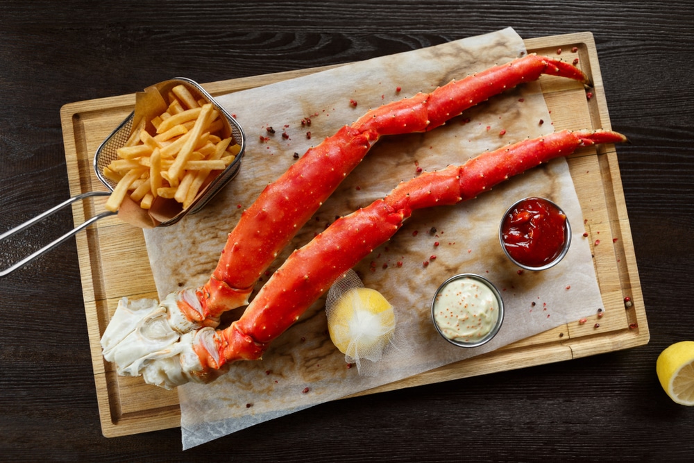 Alaskan king crab legs with butter and tomato sauces and french fries served on wooden board, top view