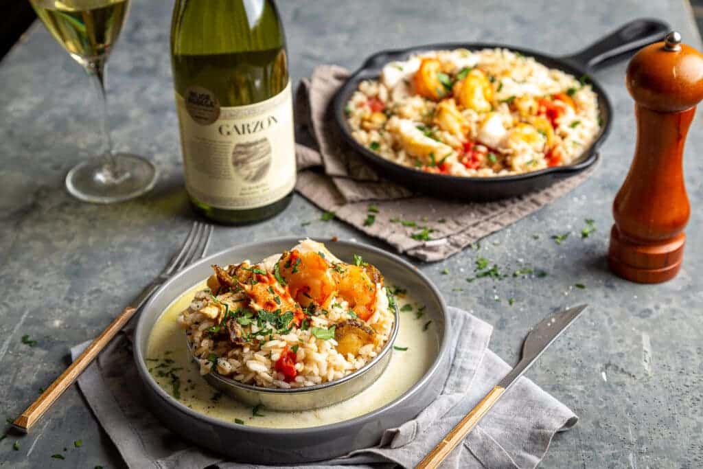 a bowl of garnished seafood risotto placed along side a bottle of wine glass fork and knife