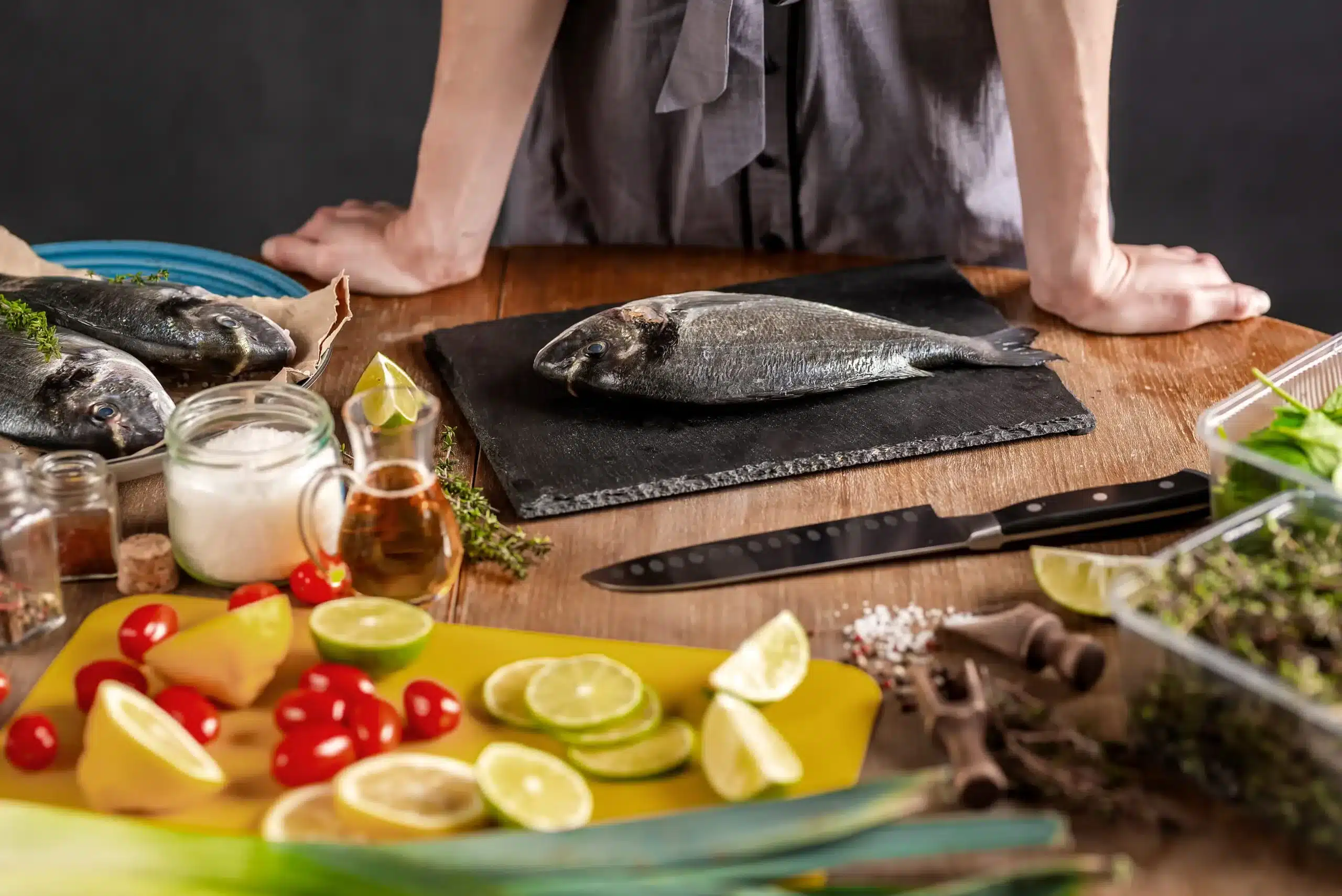 Fresh fish being prepared with herbs lemon and vegetables on a wooden kitchen counter
